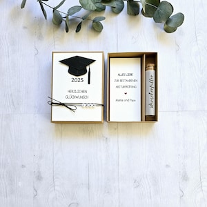 May include: A graduation gift box set with a black graduation cap, a card that says "Alles Liebe zur bestandenen Abiturpr&uuml;fung Mama und Papa" and a small glass bottle with a white label that says "w&uuml;nscheerf&uuml;ller".