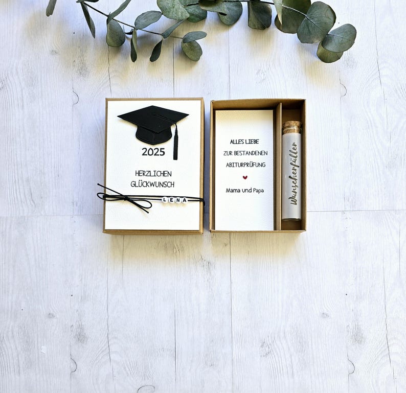 May include: A graduation gift box set with a black graduation cap, a card that says "Alles Liebe zur bestandenen Abiturpr&uuml;fung Mama und Papa" and a small glass bottle with a white label that says "w&uuml;nscheerf&uuml;ller".