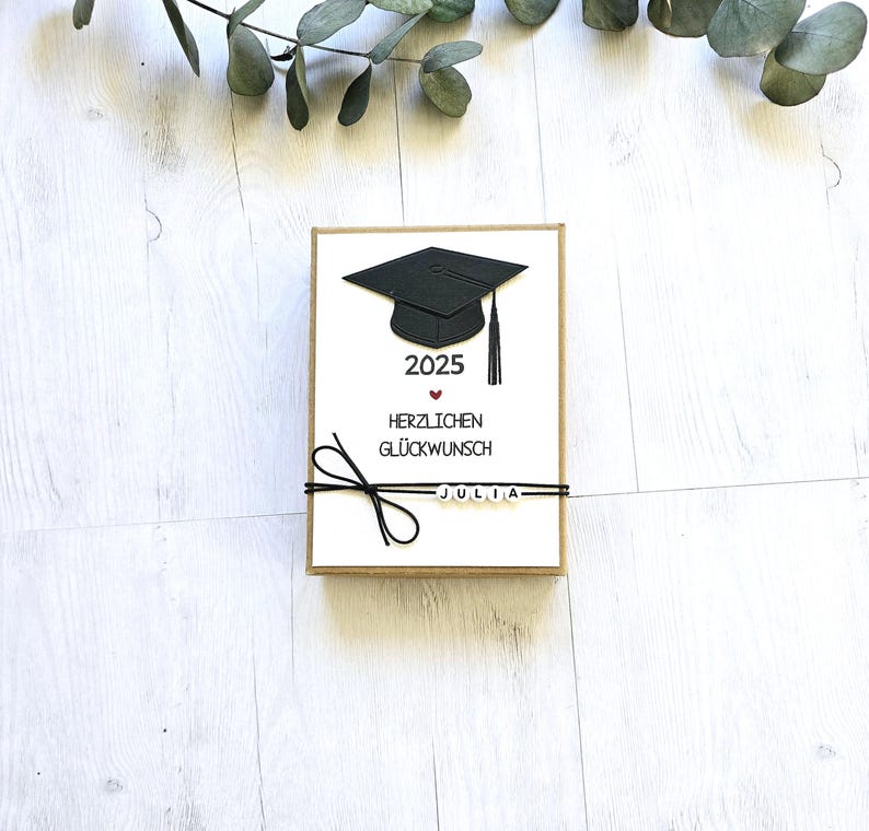 May include: A white card with a black graduation cap, the year 2025, a red heart, and the text "Herzlichen Gl&uuml;ckwunsch" in German. The card has a black ribbon tied around it with the name "Julia" spelled out in white beads.