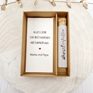 May include: A small brown box with a white card inside that reads "Alles Liebe zur bestandenen Abiturpr&uuml;fung" with a red heart and "Mama und Papa".  The box also contains a glass vial with a cork stopper and a white label that reads "Wunschersch&ouml;pfer".