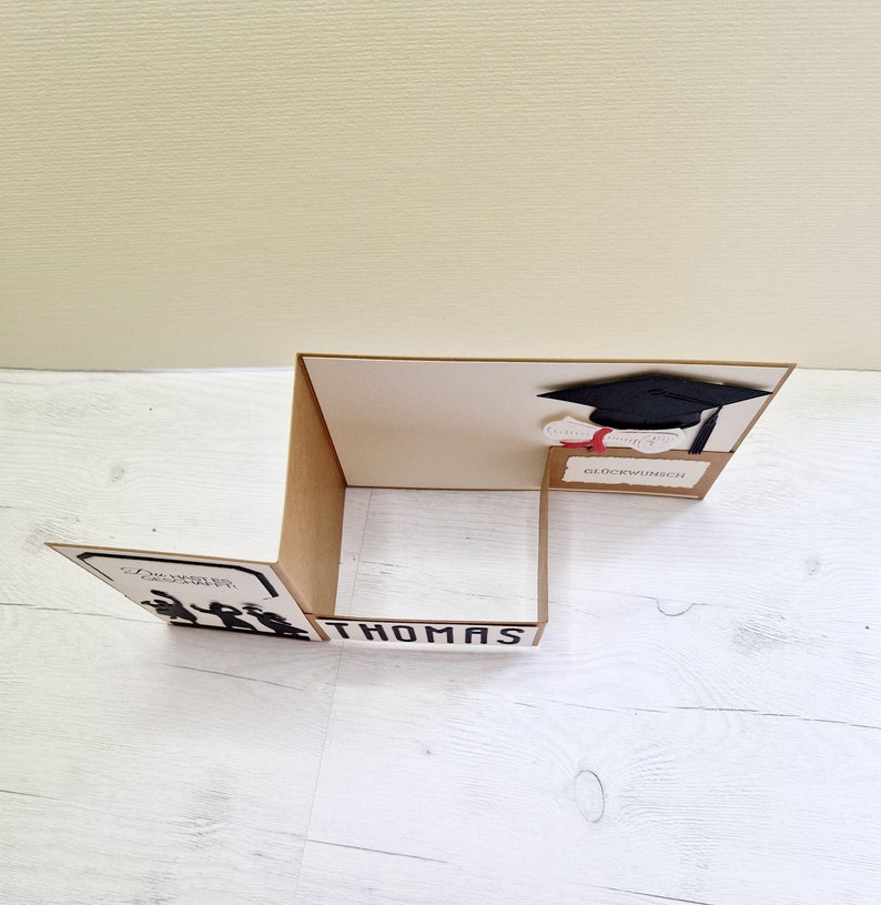 May include: A brown card with a black graduation cap and a red diploma. The card says "Gl&uuml;ckwunsch" which is German for congratulations. The card also has a silhouette of people walking in a graduation procession. The card says "Thomas" at the bottom.