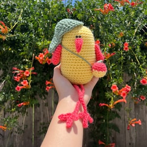 May include: A crocheted yellow bird with pink legs and a green hat. The bird is being held by a hand in front of a green bush with orange flowers.