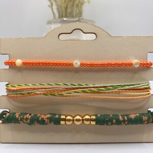 May include: Three bracelets displayed on a wooden card. The top bracelet is made of orange beads with a white bead in the center. The middle bracelet is made of multi-colored string. The bottom bracelet is made of green and brown beads with gold beads in the center.