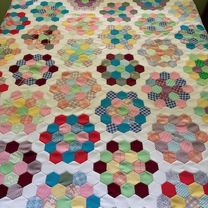 May include: A handmade quilt featuring a repeating pattern of colorful fabric hexagons arranged in floral-like clusters. The quilt is constructed with a white fabric background, showcasing a variety of solid, plaid, and floral prints.