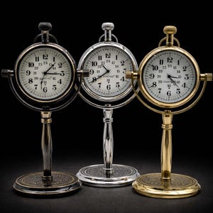 May include: Three antique-style desk clocks with white faces and black numerals. The clocks have a circular design with a stand and are in silver, bronze, and gold tones. The text "PORTHO 16" is visible on the clock faces.