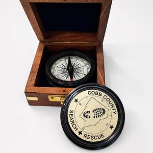 May include: A wooden box with a brass latch containing a magnetic compass. The compass lid is brass-coloured with black text reading COBB COUNTY SEARCH + RESCUE, a central boot print graphic, and cardinal directions N, S, E, and W with stars.