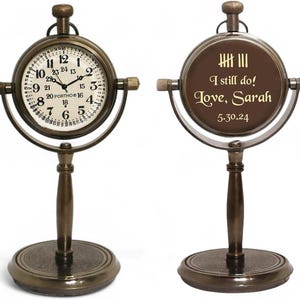 May include: A vintage-style brass desk clock with a round face and a stand. The clock face has black numerals and hands. The back of the clock has the text "I still do! Love, Sarah 5.30.24".