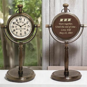 May include: A bronze-toned metal desk clock with a circular frame and a pedestal base. The clock face features black numerals and the text PORTHO. The reverse side displays the engraved text: Years together, Until the end of time, Love, Elif, May 22, 2005.