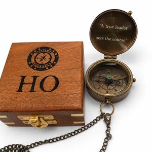 May include: A brass pocket compass with a chain and an open lid featuring the text "A true leader sets the course" next to a square wooden box engraved with "HO" and a circular "Spirit of Adventure" logo with a compass rose design.
