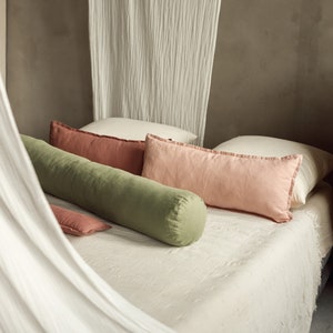 Può includere: Un letto bianco con un baldacchino bianco e una varietà di cuscini. I cuscini sono in tonalità di rosa, verde e marrone. Il letto è fatto con un lenzuolo di lino bianco.