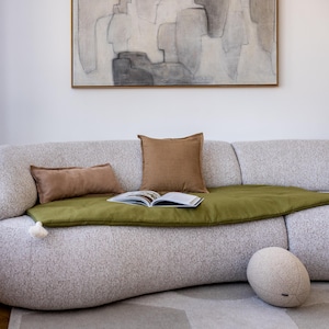 Könnte beinhalten: Ein weißes, geschwungenes Sofa mit einer grünen Wolldecke und braunen Kissen. Ein runder, beiger Hocker steht auf dem Boden vor dem Sofa.
