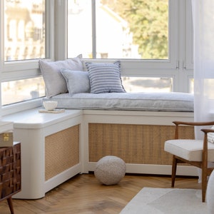 May include: A white window seat with blue and white striped cushions and pillows. The seat is in a corner of a room with a white radiator cover and a woven rattan panel. A white chair with a striped cushion sits in front of the window seat.