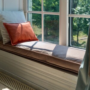 May include: A window seat with a white cushion and a coral-colored pillow. The seat is covered in a light brown fabric. A blue curtain with a tassel hangs to the right of the window.