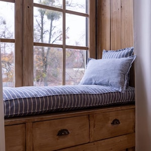 May include: A wooden window seat with a blue and white striped cushion and two drawers. The window seat is in front of a window with a view of trees.