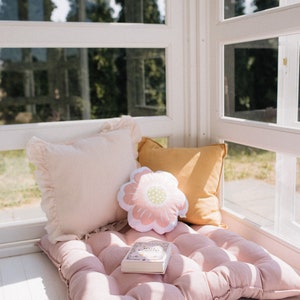 May include: A pink tufted cushion with a book and two pillows on top. One pillow is pink with a ruffled edge and the other is yellow. A flower-shaped pillow is in the center. The cushion is in a sunlit room with a window behind it.