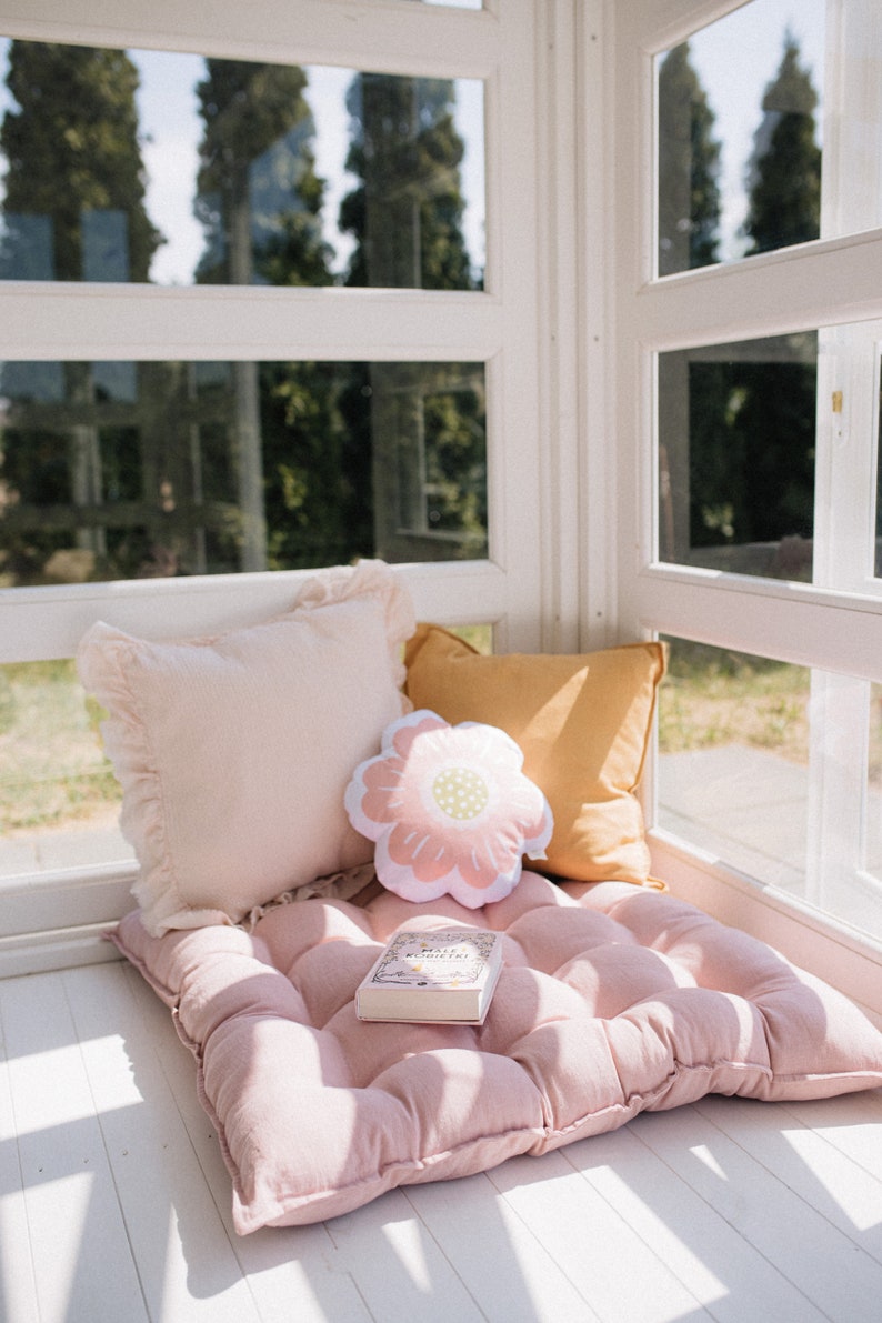 May include: A pink tufted cushion with a book and two pillows on top. One pillow is pink with a ruffled edge and the other is yellow. A flower-shaped pillow is in the center. The cushion is in a sunlit room with a window behind it.