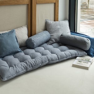 May include: A gray, tufted floor cushion with a variety of pillows in shades of blue and gray. The cushion is placed in front of a window with a view of a city street.
