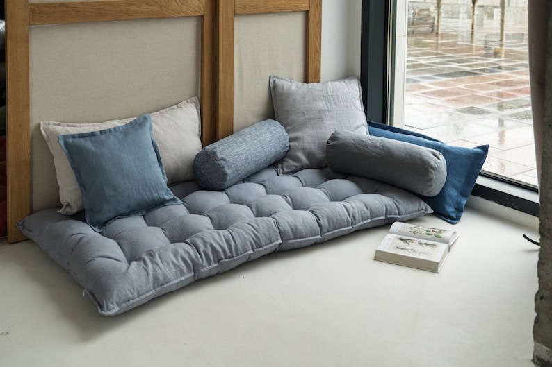 May include: A gray, tufted floor cushion with a variety of pillows in shades of blue and gray. The cushion is placed in front of a window with a view of a city street.