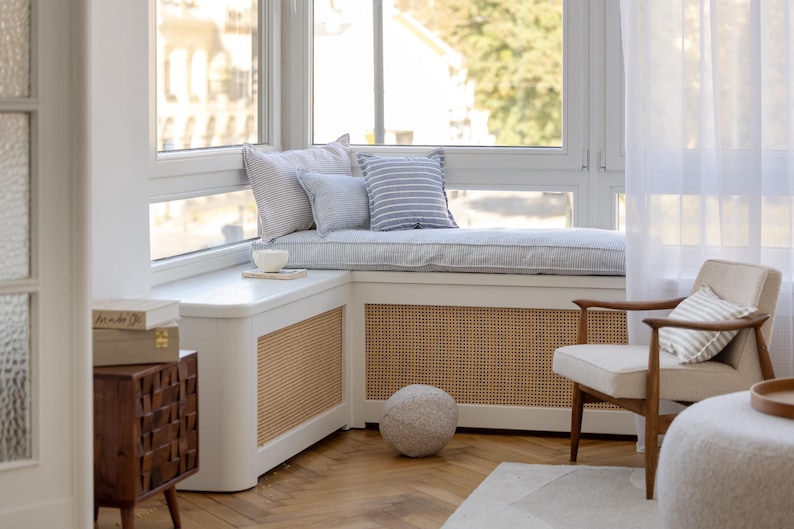 May include: A white window seat with blue and white striped cushions and pillows. The seat is in a corner of a room with a white radiator cover and a woven rattan panel. A white chair with a striped cushion sits in front of the window seat.