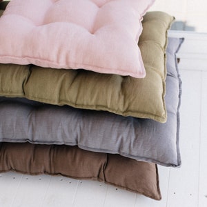 May include: A stack of four square floor cushions in different colors. The top cushion is pink, the next is olive green, the third is gray, and the bottom is brown.