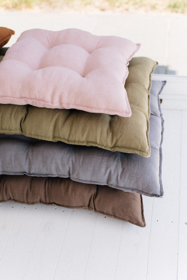 May include: A stack of four square floor cushions in different colors. The top cushion is pink, the next is olive green, the third is gray, and the bottom is brown.