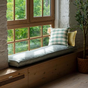 May include: A window seat featuring a light blue cushion and two pillows. One pillow displays a green and white check pattern, the other has yellow and white stripes. The window frame is wooden.