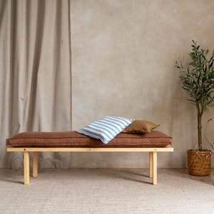 May include: A wooden bench with a rust-colored cushion and two pillows. One pillow has blue and white stripes, and the other is brown. The bench has a natural wood frame and is set against a neutral-toned wall.