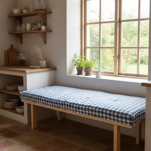 Peut inclure: Un banc en bois doté d'un coussin rectangulaire à motif vichy bleu et blanc. Le banc possède quatre pieds droits et est placé devant une fenêtre. Le coussin est capitonné avec des indentations visibles sur toute sa surface.
