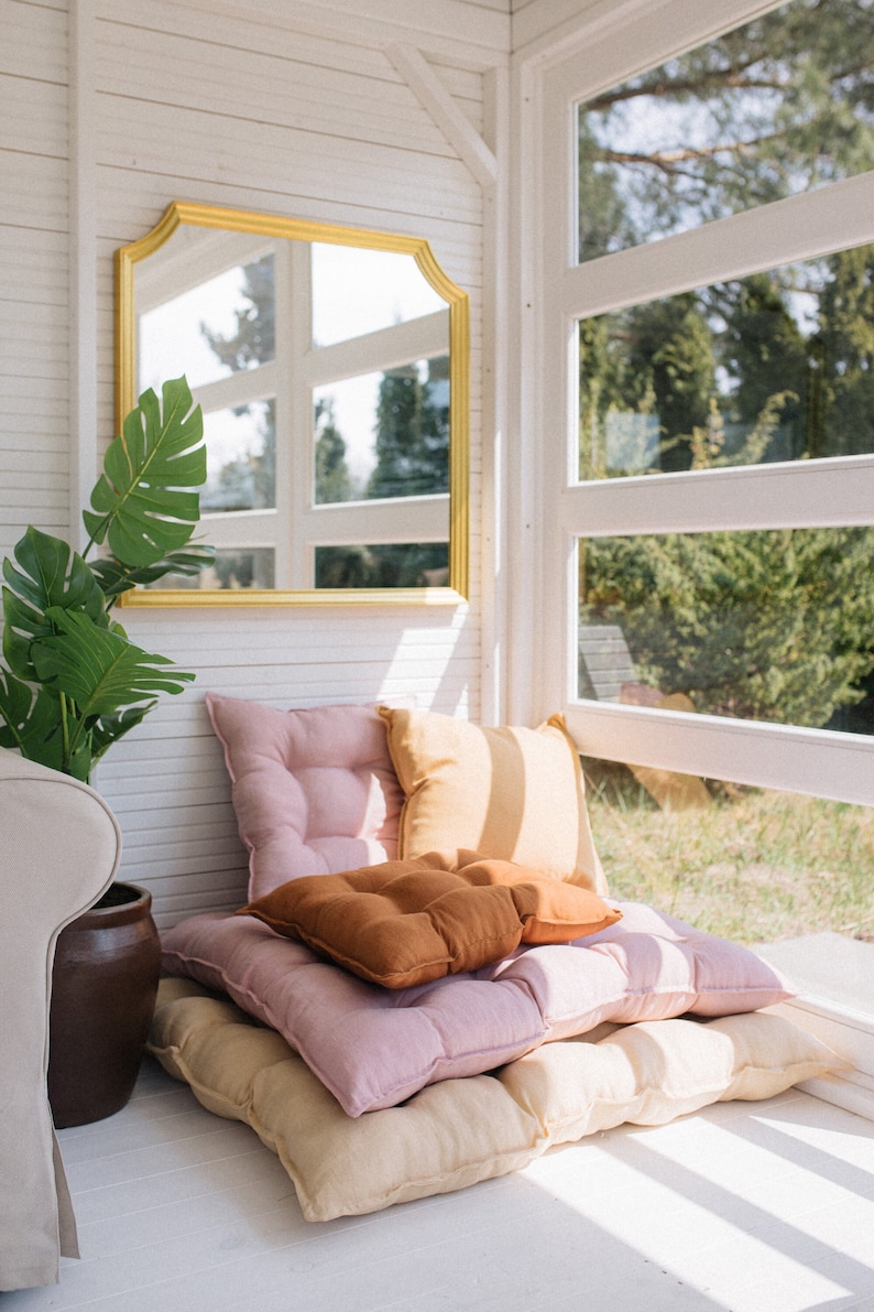 May include: A white room with a large window and a gold framed mirror. There are three cushions on the floor, one pink, one beige, and one brown.