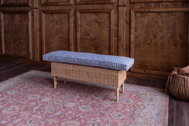 May include: A woven storage bench with a blue and white striped cushion. The bench is sitting on a red patterned rug in front of a wood paneled wall.