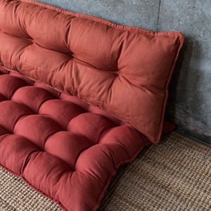 May include: Two red, tufted floor cushions made of linen fabric. The cushions are stacked on top of each other and are sitting on a woven rug.