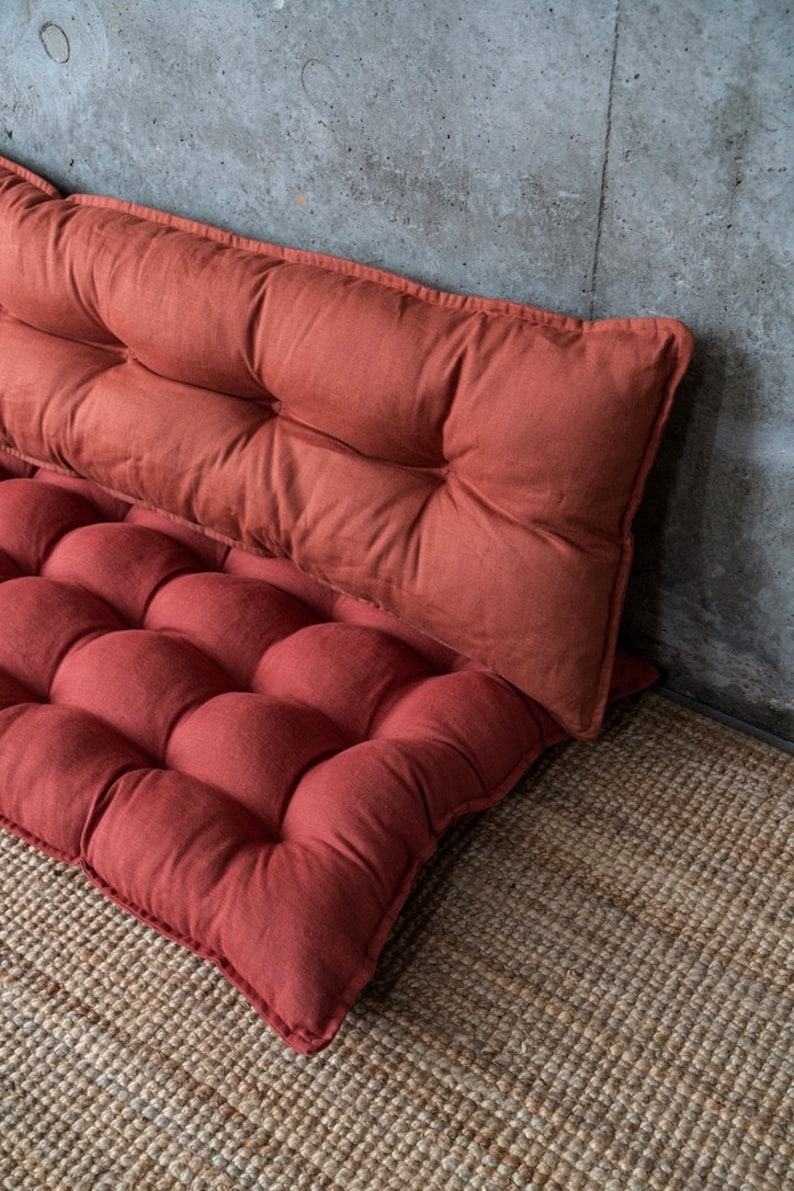 May include: Two red, tufted floor cushions made of linen fabric. The cushions are stacked on top of each other and are sitting on a woven rug.