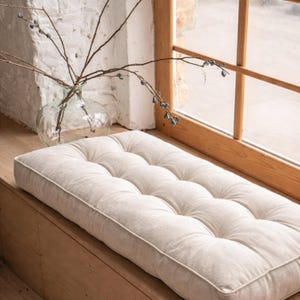 Peut inclure: Un coussin rectangulaire capitonné de couleur beige neutre. Le coussin est posé sur une surface en bois près d'une fenêtre. Un vase en verre transparent avec des branches est visible en arrière-plan. Le coussin mesure environ 76 cm de long.