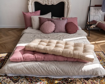 May include: A bed with a white linen duvet and two pink and beige cushions. The bed is on a patterned rug. There are several pillows on the bed, including two round pillows, one with a pink and white pattern and the other with a burgundy color.