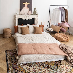 Könnte beinhalten: Ein weißes Bett mit einem beigefarbenen Bettbezug und einer braunen Wolldecke. Das Bett ist mit mehreren Kissen dekoriert, darunter braune, beige und weiße. Das Bett steht in einem Raum mit einer weißen Wand und einem Kamin. Vor dem Bett liegt ein Teppich auf dem Boden.