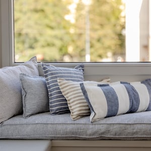 May include: A window seat with a striped cushion and several blue and white striped pillows. The window seat is positioned in front of a window with a view of trees.