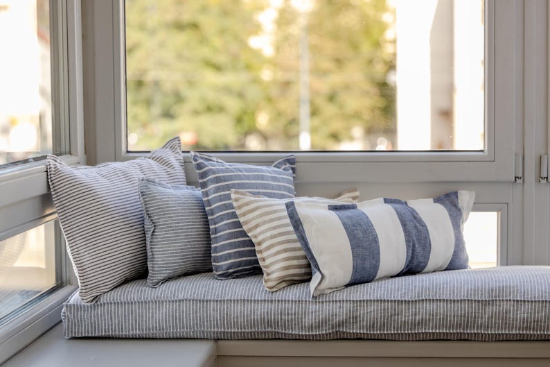 May include: A window seat with a striped cushion and several blue and white striped pillows. The window seat is positioned in front of a window with a view of trees.