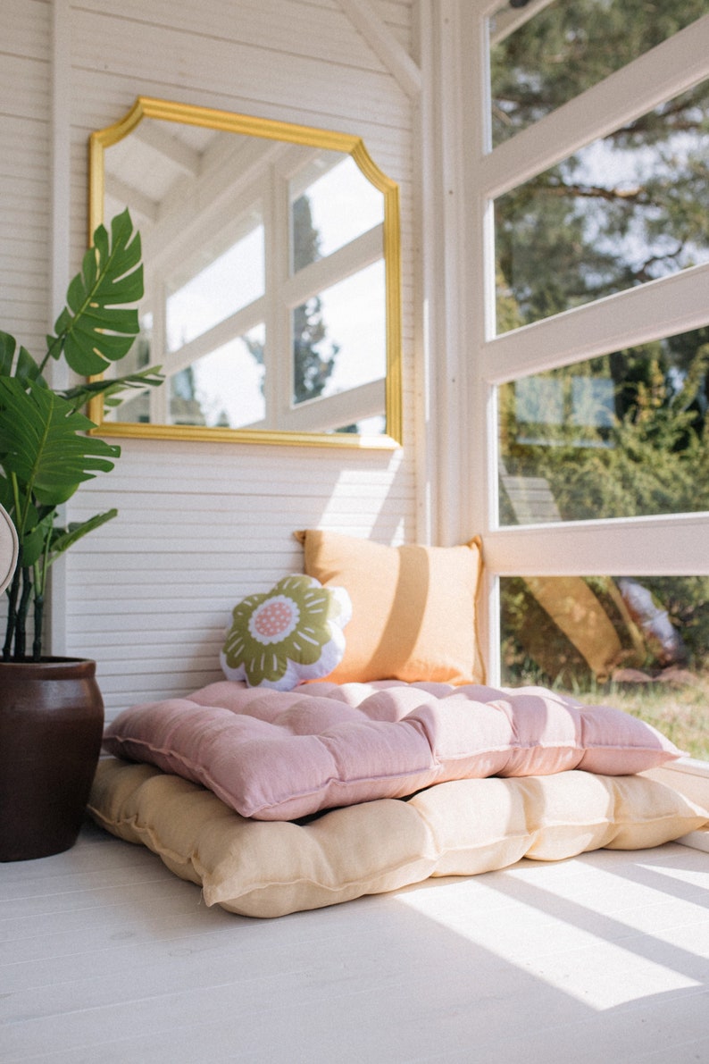 May include: A pink and beige floor cushion with a floral pillow on top. The cushion is in a sunlit room with a large window and a gold framed mirror on the wall.