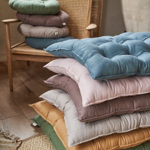 May include: A stack of seven floor cushions in various colors, including green, brown, beige, and blue. The cushions are made of a soft, textured fabric and are perfect for adding comfort and style to any room.