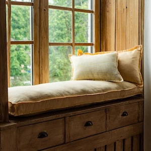 May include: A cozy window seat with a cream-colored cushion and two pillows. The pillows are light yellow with a striped pattern. The window frame and the cabinet below are made of wood. The window overlooks a green, leafy view.