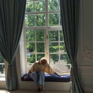 Op de afbeelding: Een persoon zit op een vensterbank in een kamer met grote ramen en blauwe gordijnen. De persoon draagt een geel shirt en een blauwe jeans. Het raam is open en er zijn bomen buiten.