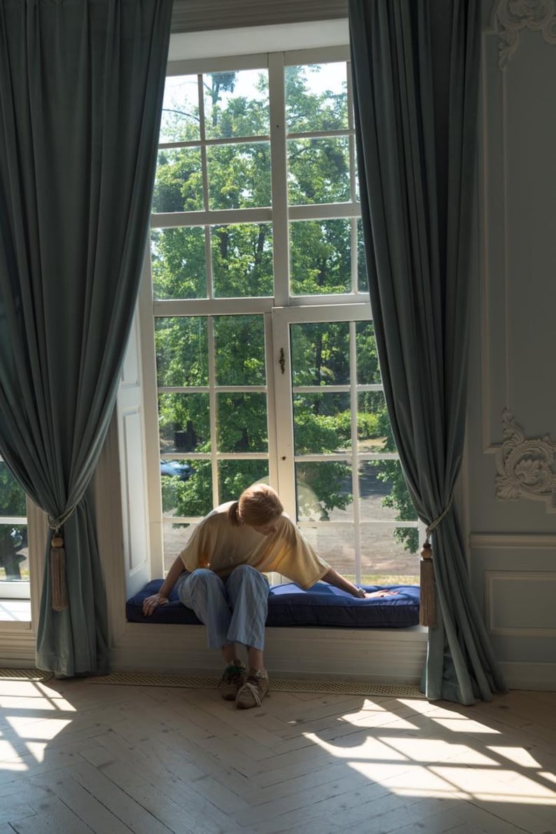 Op de afbeelding: Een persoon zit op een vensterbank in een kamer met grote ramen en blauwe gordijnen. De persoon draagt een geel shirt en een blauwe jeans. Het raam is open en er zijn bomen buiten.
