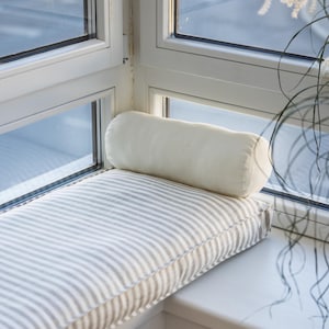May include: A white window seat cushion with gray stripes and a white bolster pillow. The cushion is placed in a corner window seat with white trim and a wicker-style radiator cover.