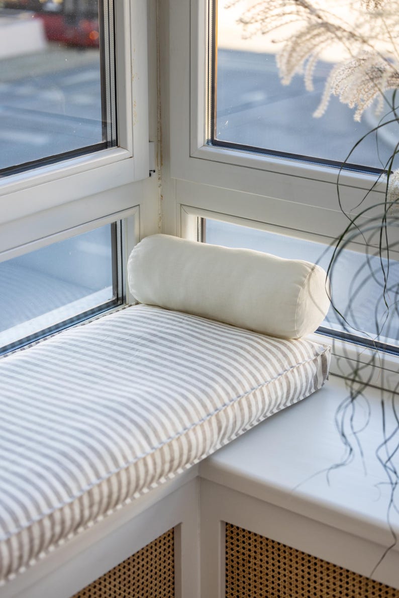 May include: A white window seat cushion with gray stripes and a white bolster pillow. The cushion is placed in a corner window seat with white trim and a wicker-style radiator cover.
