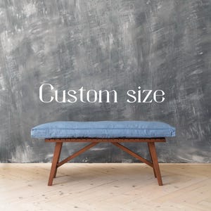 May include: A wooden bench with a blue linen cushion. The bench has a simple design with a rectangular seat and four legs. The text "Custom size" is written on the wall behind the bench.
