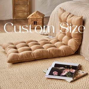 May include: A tan floor cushion with a tufted design. The cushion is on a woven rug with a diamond pattern. The text "Custom Size" is visible in the image.