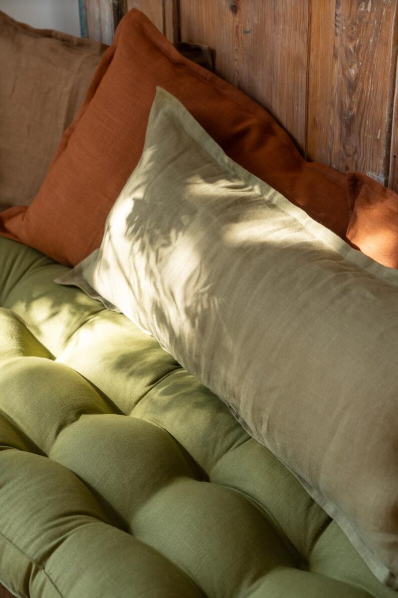 K&ouml;nnte beinhalten: Zwei Kissen aus Leinen, eines in einem verbrannten Orangeton und eines in einem hellgr&uuml;nen Ton, liegen auf einem gr&uuml;nen Leinen-Sofa.