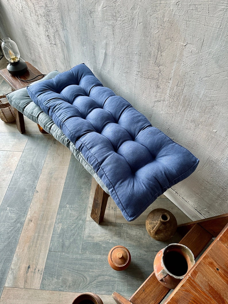 K&ouml;nnte beinhalten: Zwei blaue und graue Knopf-Steppkissen auf einer Holzbank. Die Kissen sind aus einem weichen, strukturierten Stoff gefertigt und haben ein Knopf-Steppdesign.