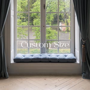 Peut inclure: Un coussin de fenêtre bleu avec des détails capitonnés est placé sur un rebord de fenêtre avec une bordure blanche. La fenêtre est ouverte et offre une vue sur des arbres verts à l'extérieur. Le texte "Custom Size" est visible dans la fenêtre.