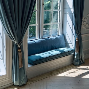 Könnte beinhalten: Ein Fensterplatz mit einem blauen Kissen und blauen Samtvorhängen mit Quasten. Das Fenster bietet einen Blick auf Bäume und einen weißen Holzrahmen.